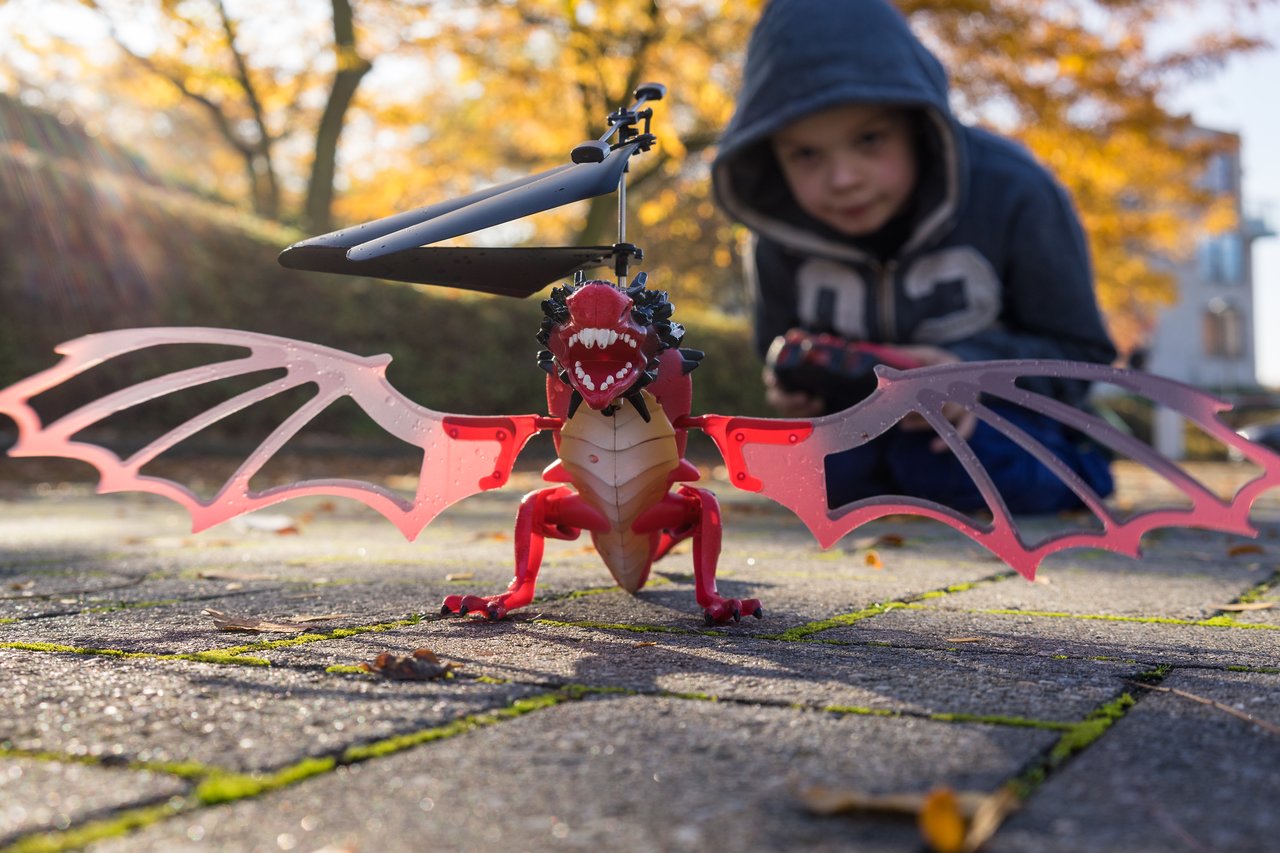 A red dragon-shaped toy helicopter with spinning blades is on the ground, while a child controls it with a remote.