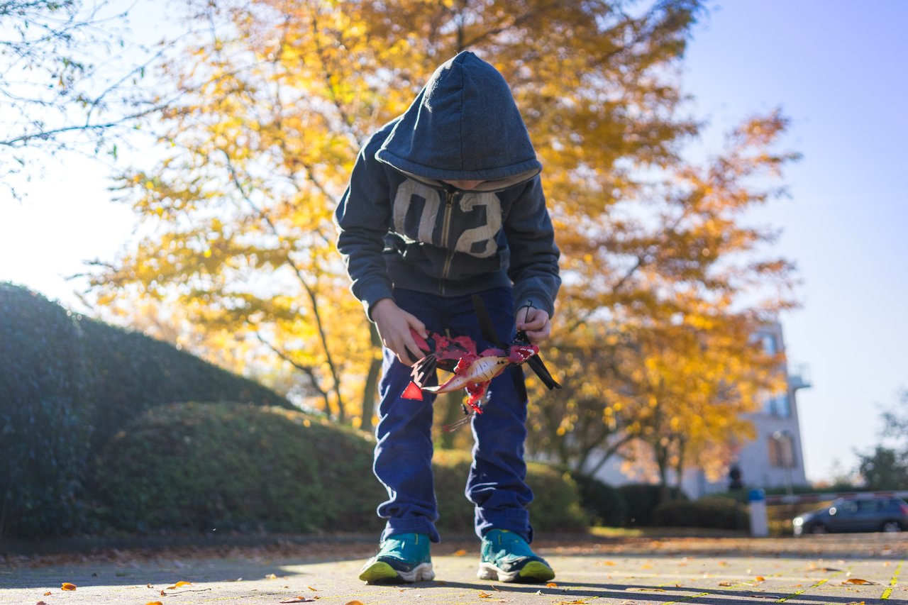 A child in a hooded jacket looks down while holding a red dragon-shaped toy outdoors on a sunny day.