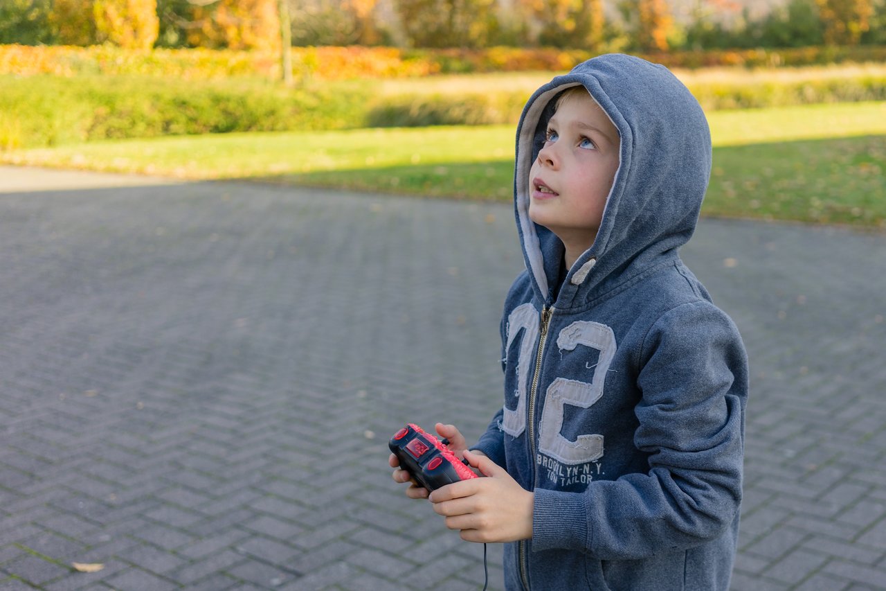 A child in a hoodie holds a remote control and looks up, possibly operating a flying toy or drone.