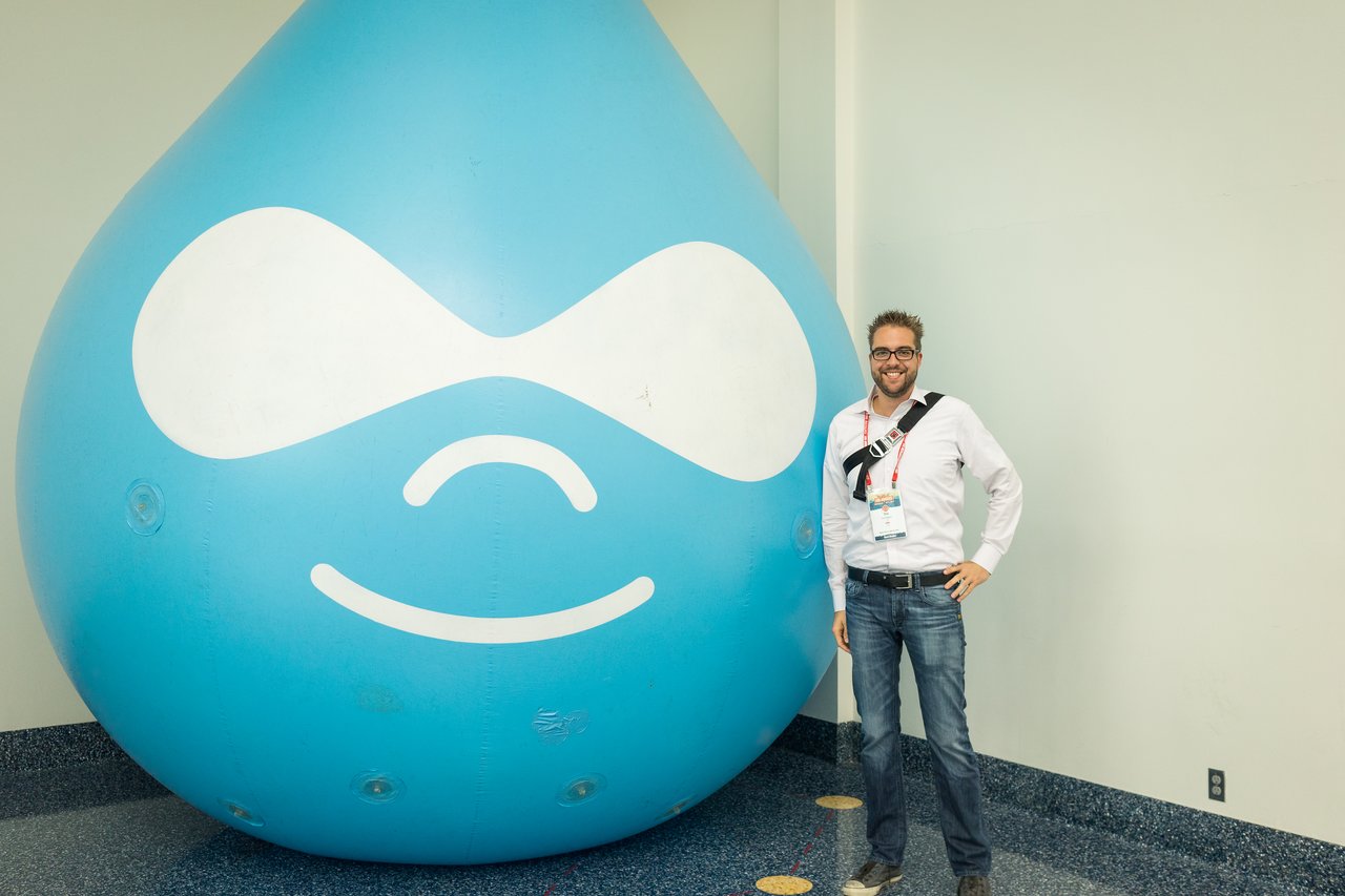 A man smiles while posing next to a large inflatable Druplicon, the Drupal logo, in an indoor setting.
