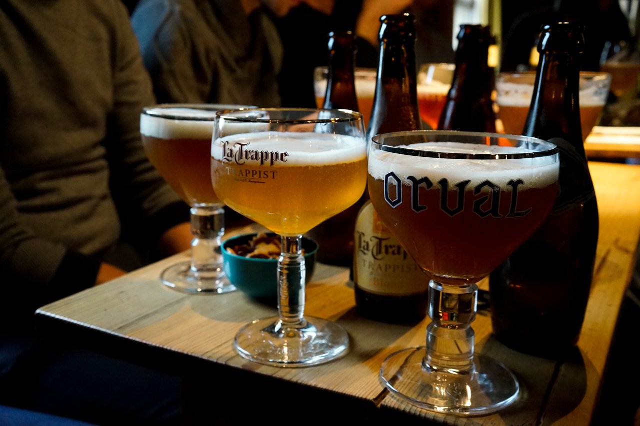 Glasses of Belgian beer on a wooden table, with bottles and a bowl of snacks in the background.