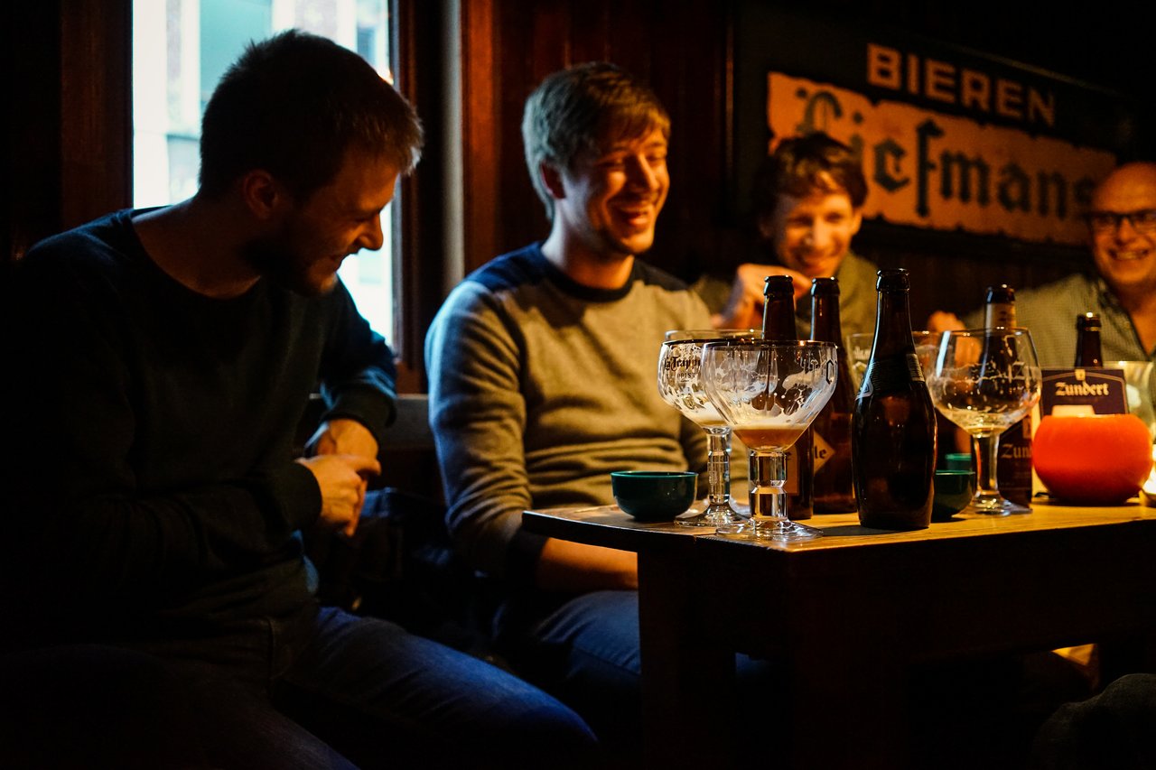 A group of friends laughs and talks at a bar table with beer bottles and glasses.