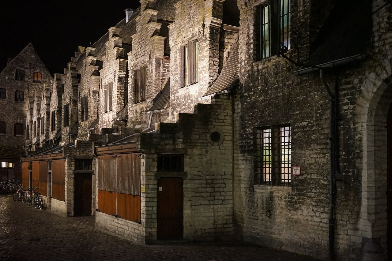 A historic brick building in Ghent at night, with stepped gables and wooden shutters, illuminated by warm lighting.