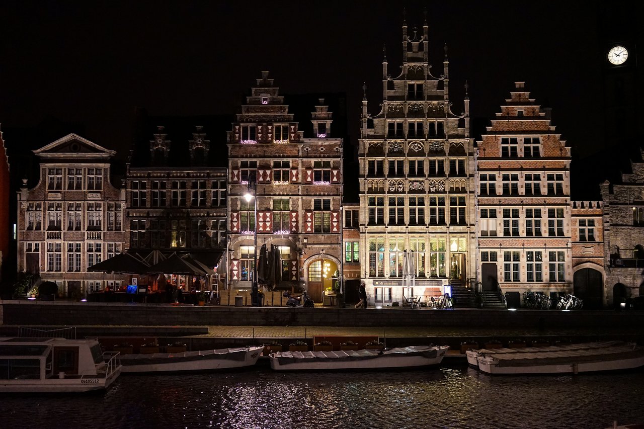 Historic buildings in Ghent illuminated at night, with boats docked along the canal in the foreground.