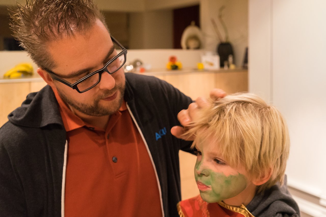 A man adjusts a child's hair while the child wears a costume and green face paint for Halloween.