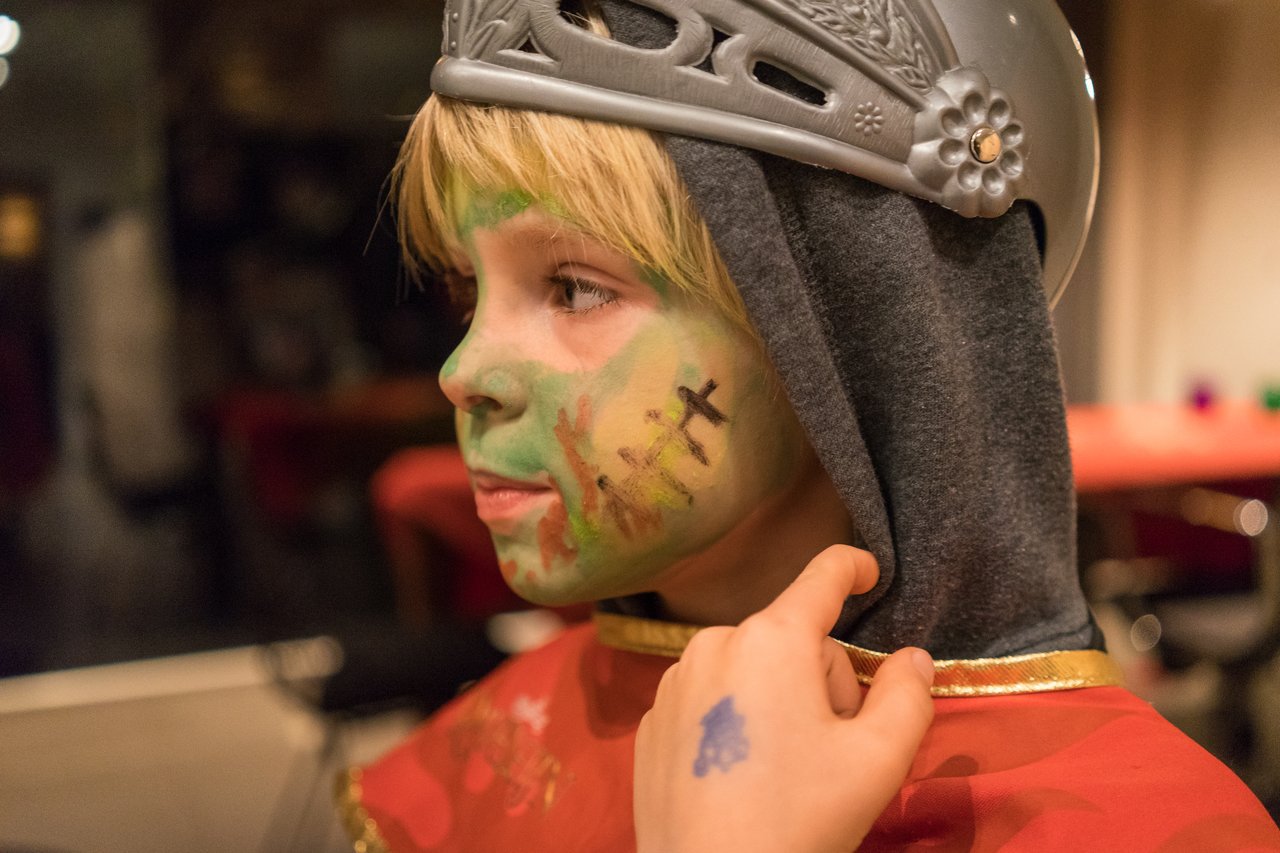 A child in a knight costume with face paint looks to the side while someone adjusts their hood.