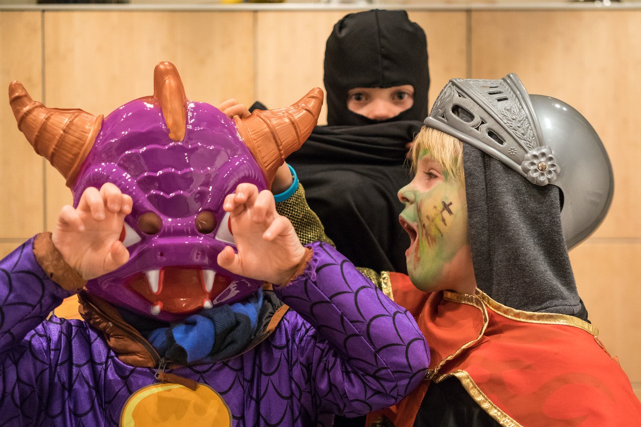 Three children in Halloween costumes pose playfully–one as a dragon, another as a knight, and one as a ninja.