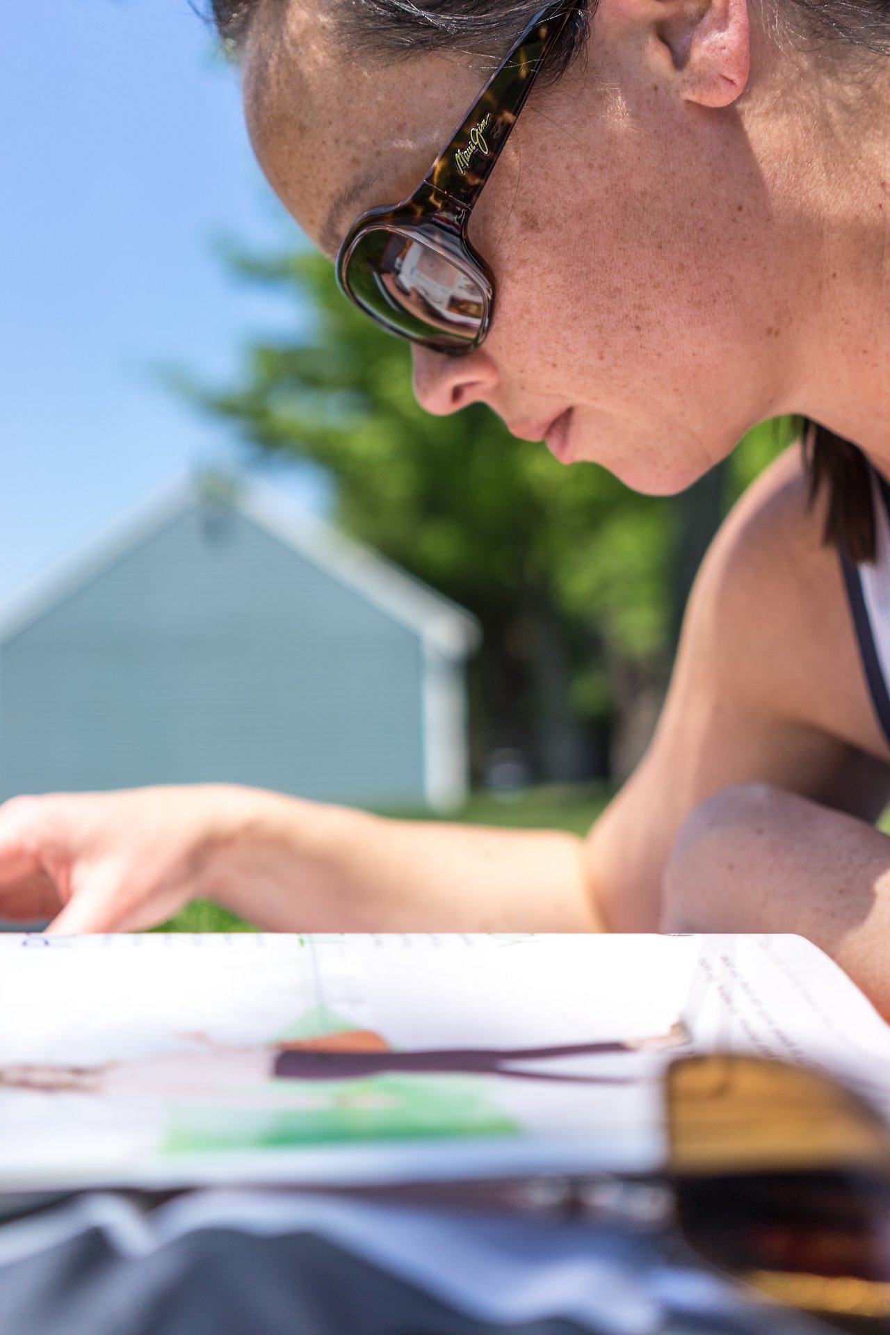 A person wearing sunglasses reads in a notebook while sitting outside in the park.