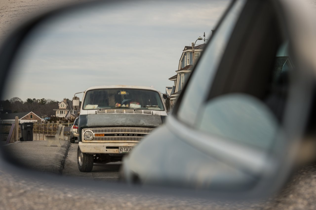 A vintage Dodge van is visible in the car's side mirror, parked on a coastal road near houses.