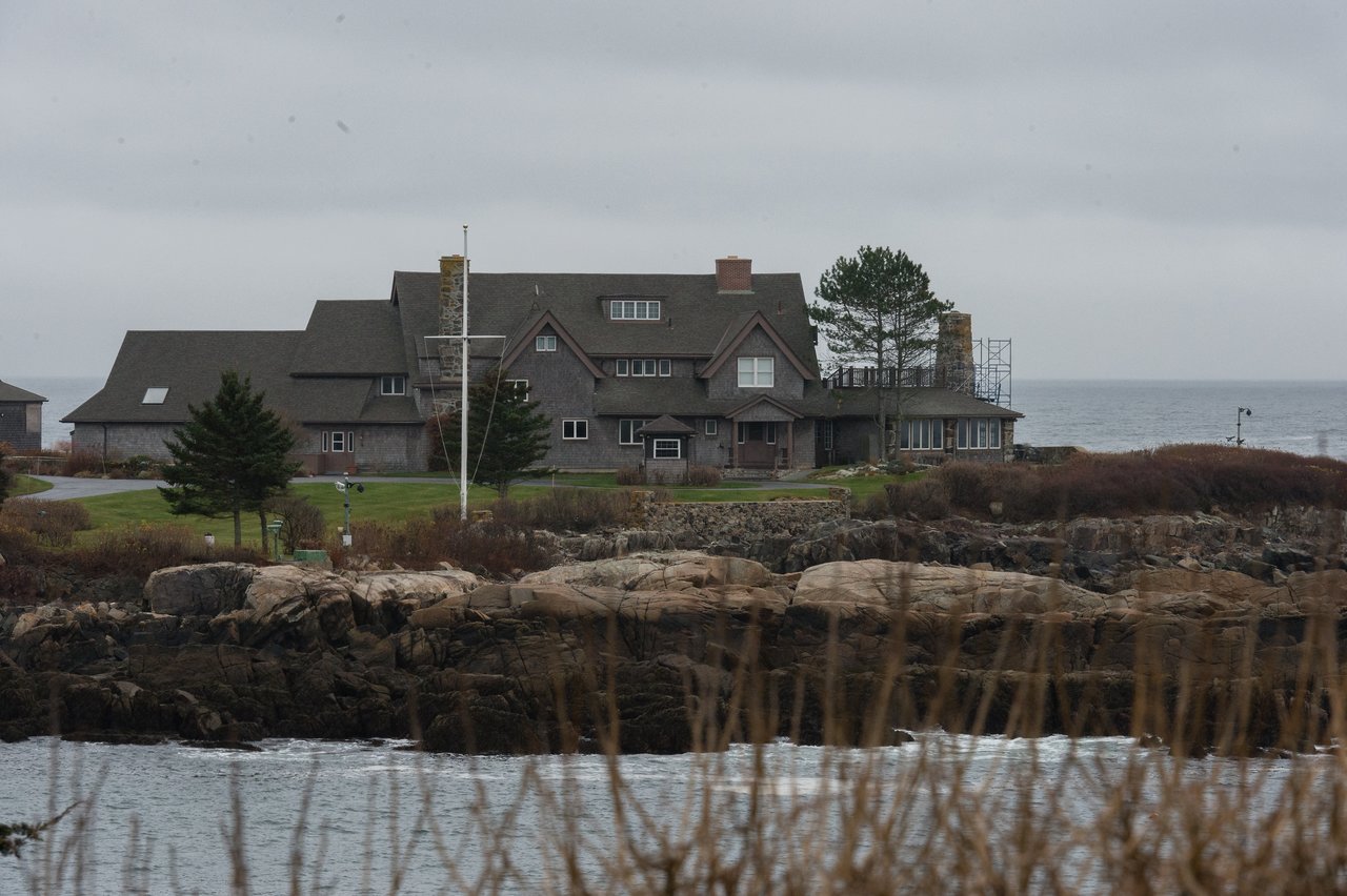 A large coastal house sits on a rocky shoreline with the ocean in the background under an overcast sky.