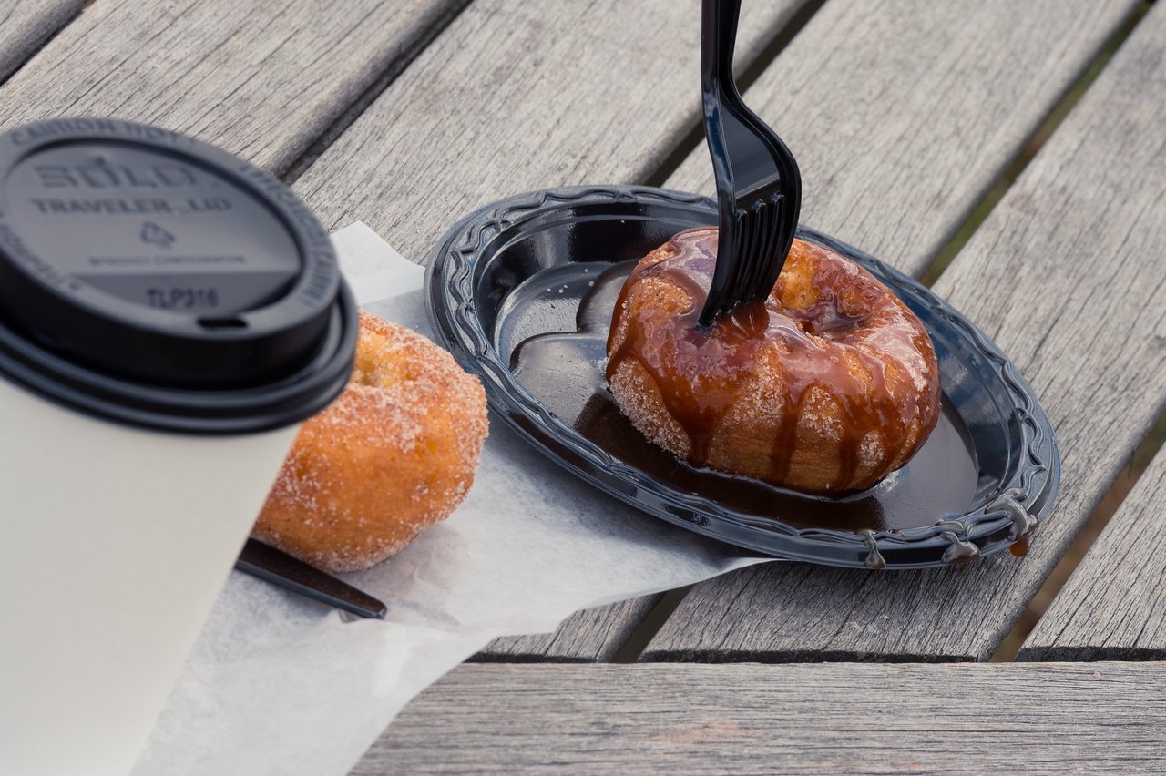 A caramel-drizzled donut with a plastic fork on a plate, next to a sugar-coated donut and a coffee cup.