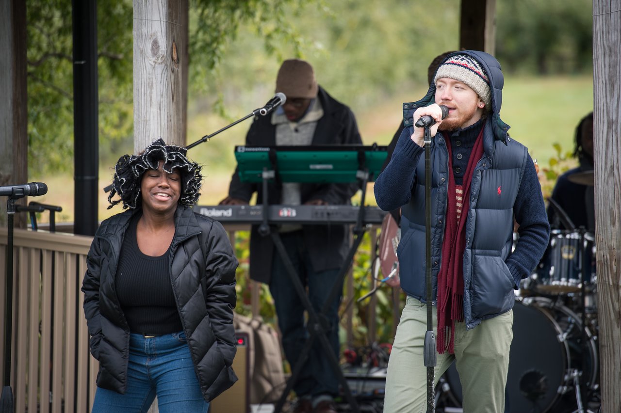 A band performs outdoors; a man sings into a microphone while a woman smiles and a keyboardist plays behind them.