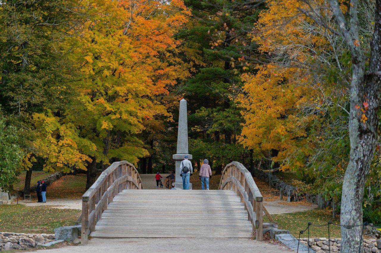 A wooden bridge with people walking toward a stone monument, surrounded by trees with autumn-colored leaves.