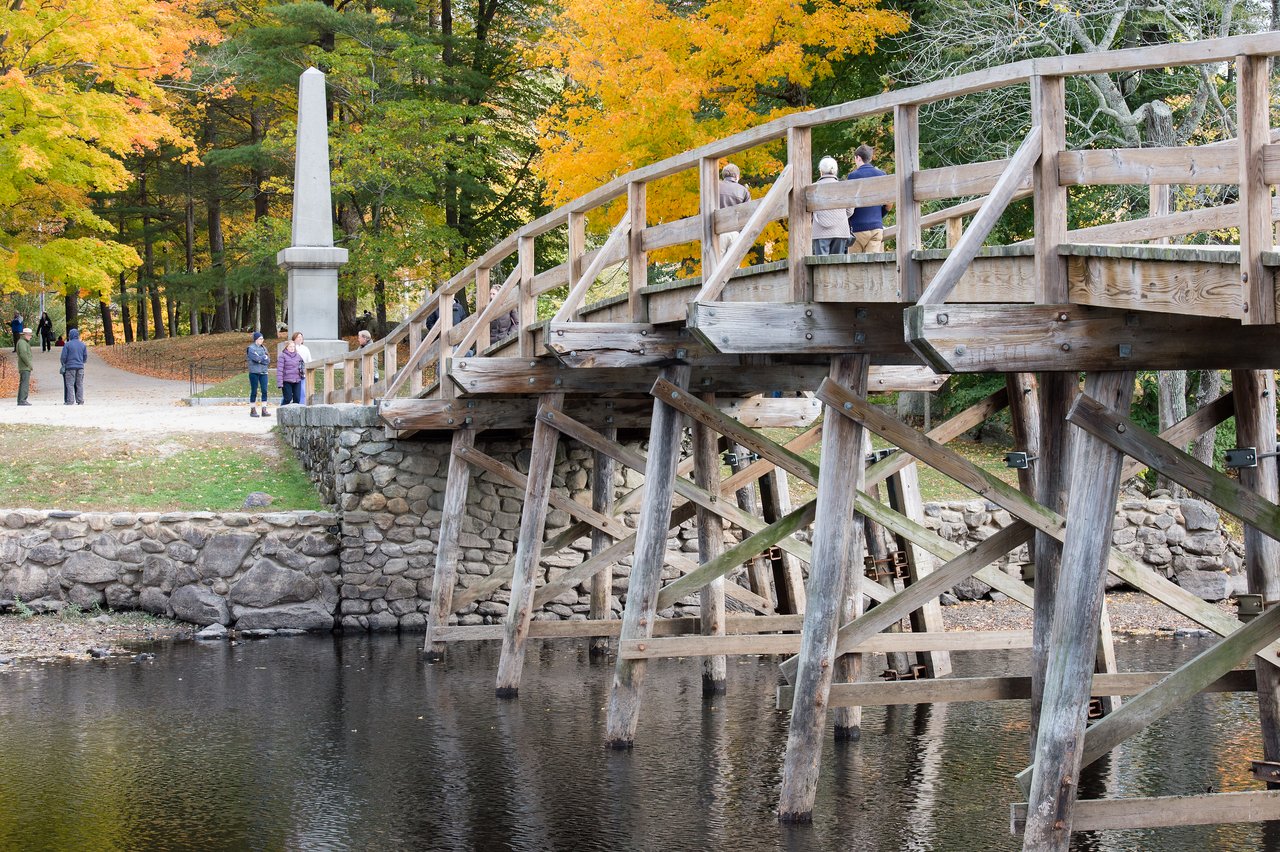 A wooden bridge spans a calm river, with people walking and standing.
