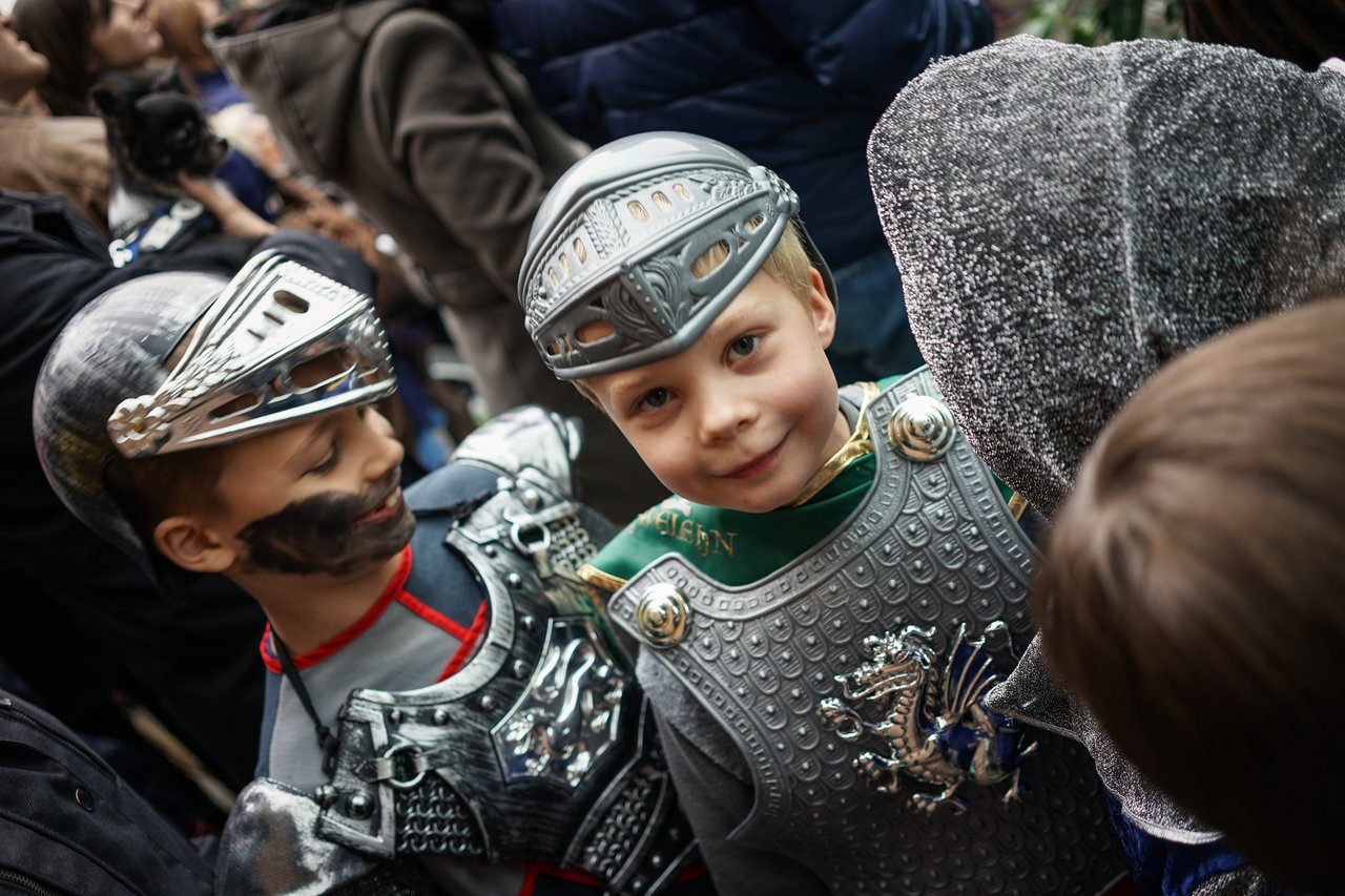 Children dressed as knights participate in a school performance, wearing armor and helmets while interacting with others in the crowd.