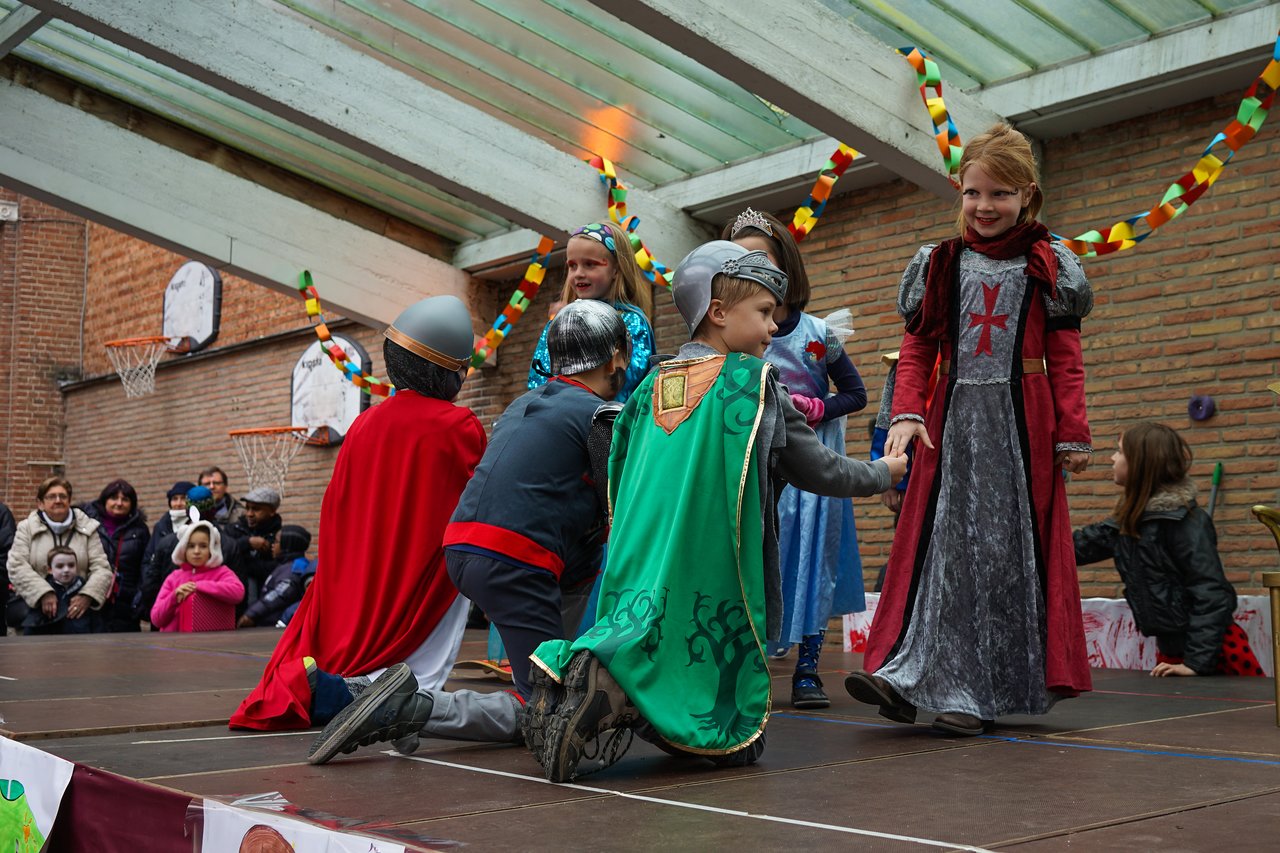 Children in medieval costumes perform on a school stage, with one kneeling and holding hands with another.