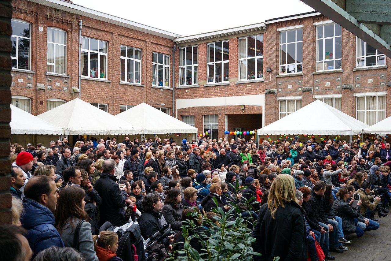 A large crowd of people sits and stands in a school courtyard, attentively watching a performance.