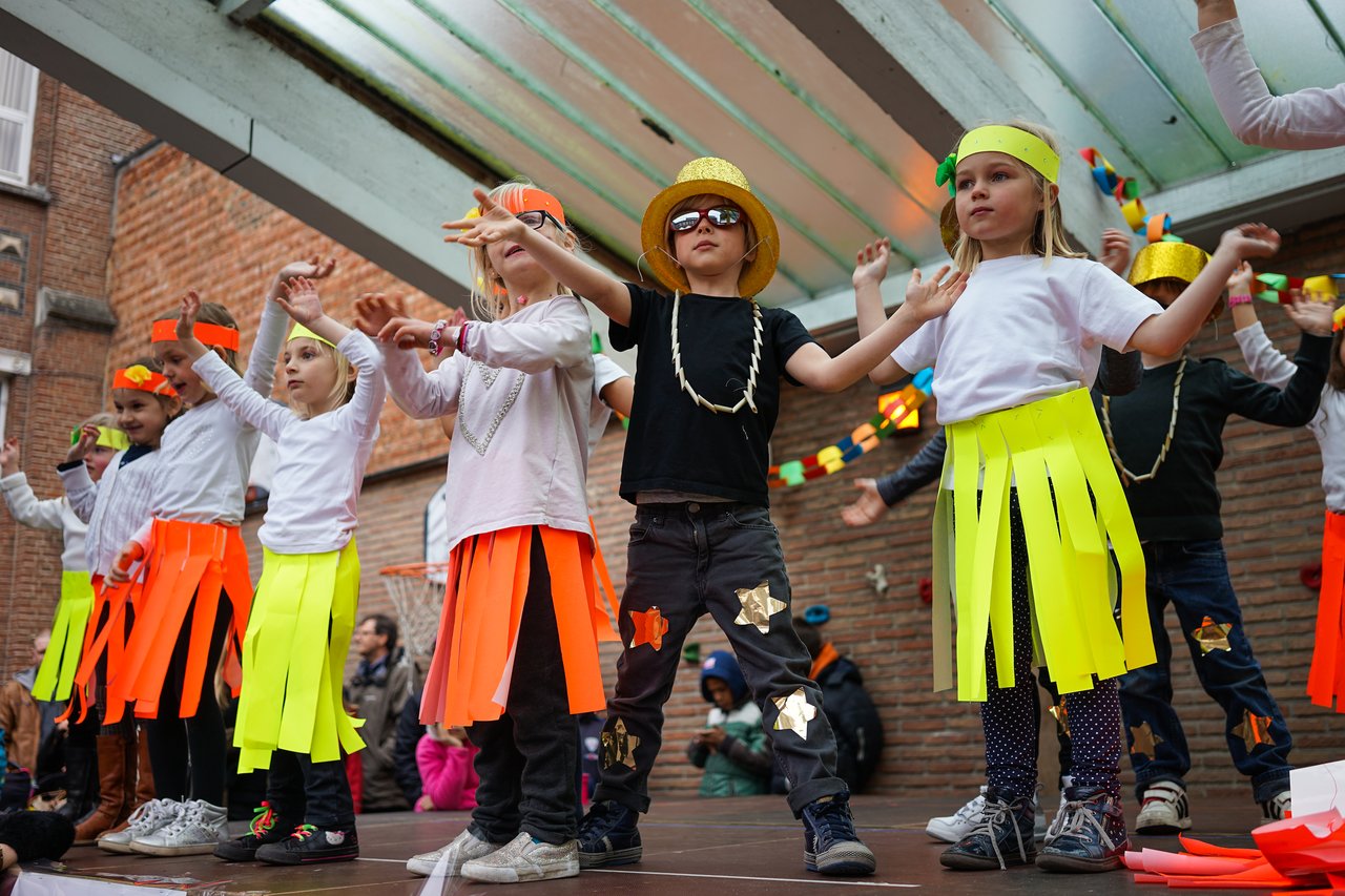 Children in colorful costumes perform a choreographed dance on an outdoor stage during a school event.