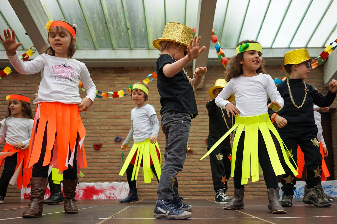 Children in colorful costumes dance on stage during a school performance, wearing paper skirts and gold hats.