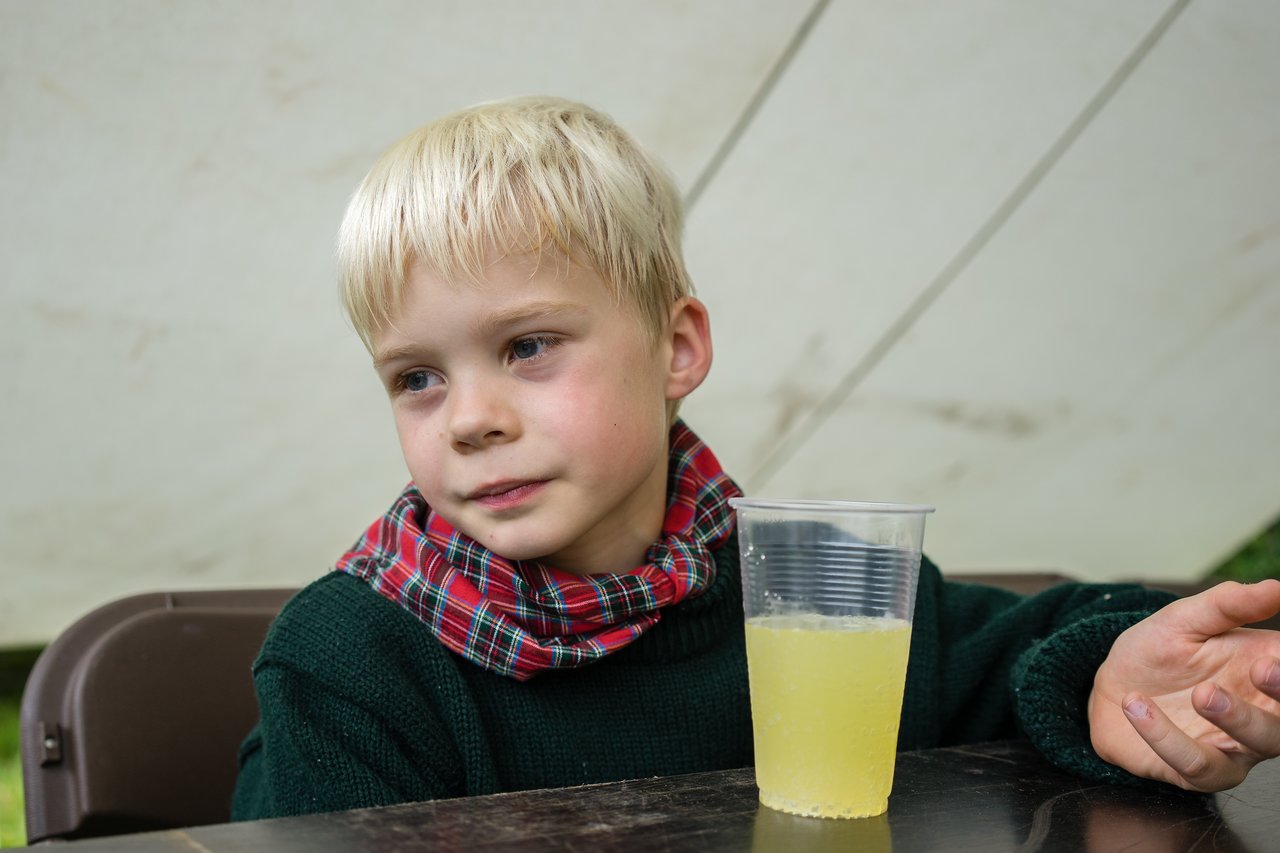 A young child in a scout uniform sits at a table with a cup of yellow drink, looking thoughtful.