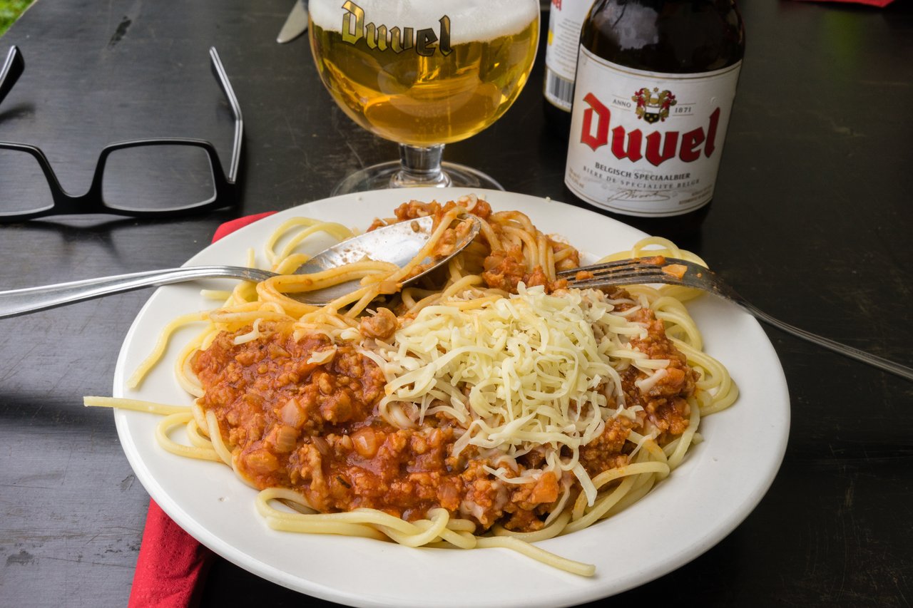 A plate of spaghetti with meat sauce and shredded cheese, with beer and glasses on a dark table.