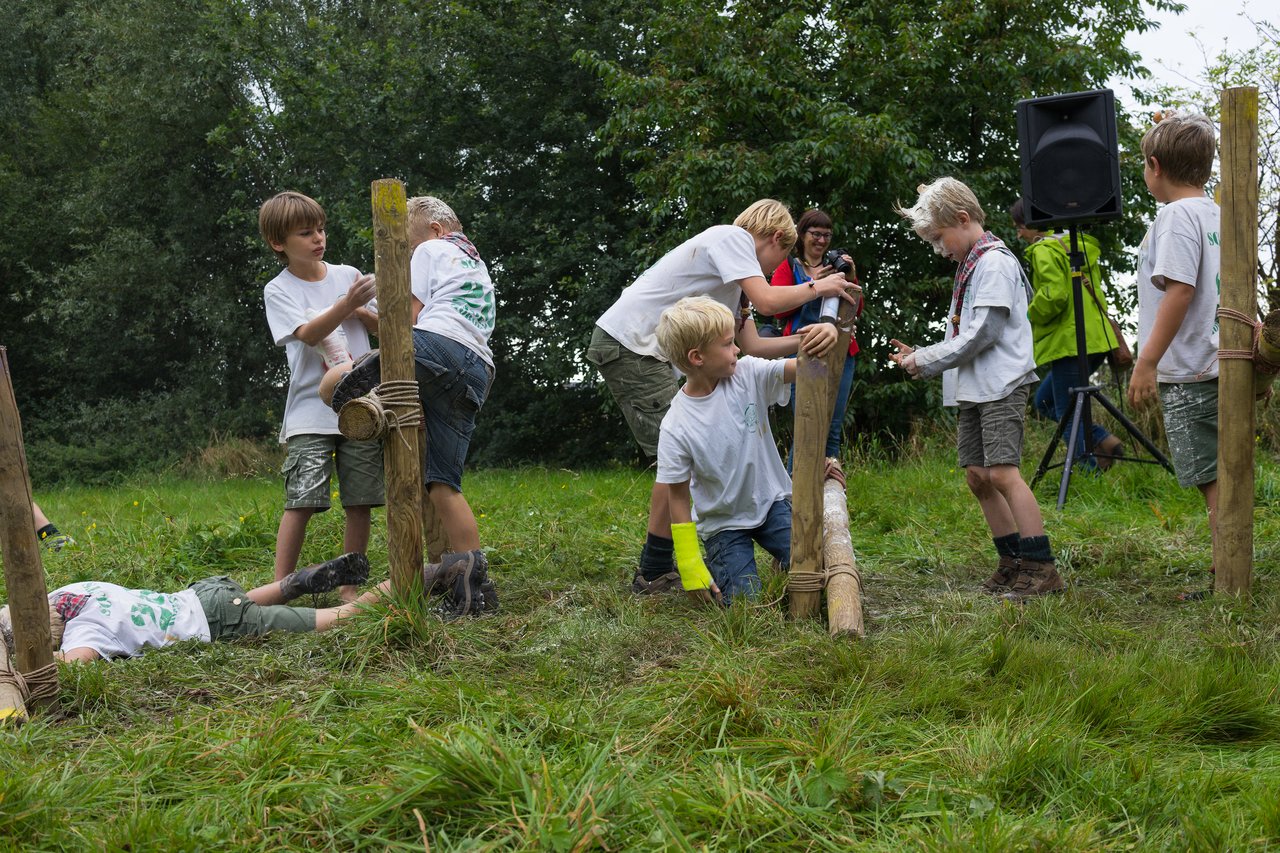 Children in scout uniforms work together to climb and cross a wooden obstacle in a grassy outdoor setting.