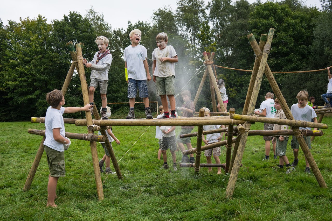 Scouts participate in an outdoor activity, crossing a wooden structure while being sprayed with water or powder.