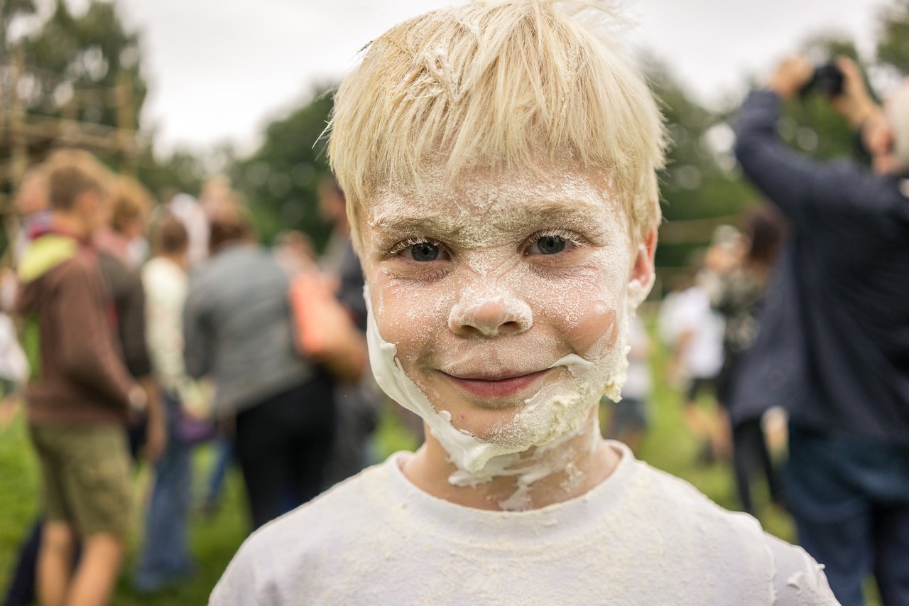A smiling child with a face covered in white powder stands outdoors during a scouting event with people in the background.
