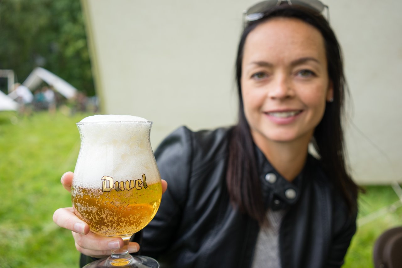 A woman in a black jacket holds a glass of beer with foam on top, offering it forward.
