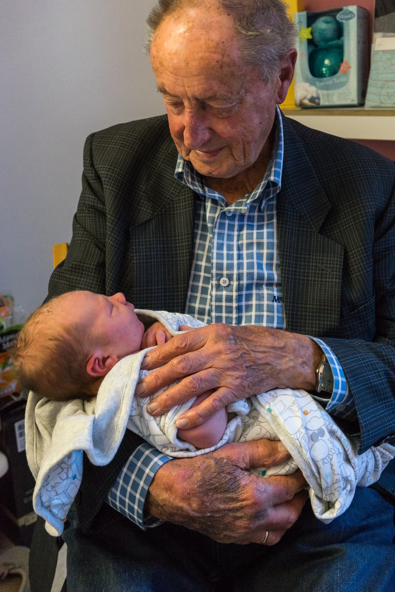 An elderly man in a checkered shirt holds a newborn baby wrapped in a blanket, looking down at them.