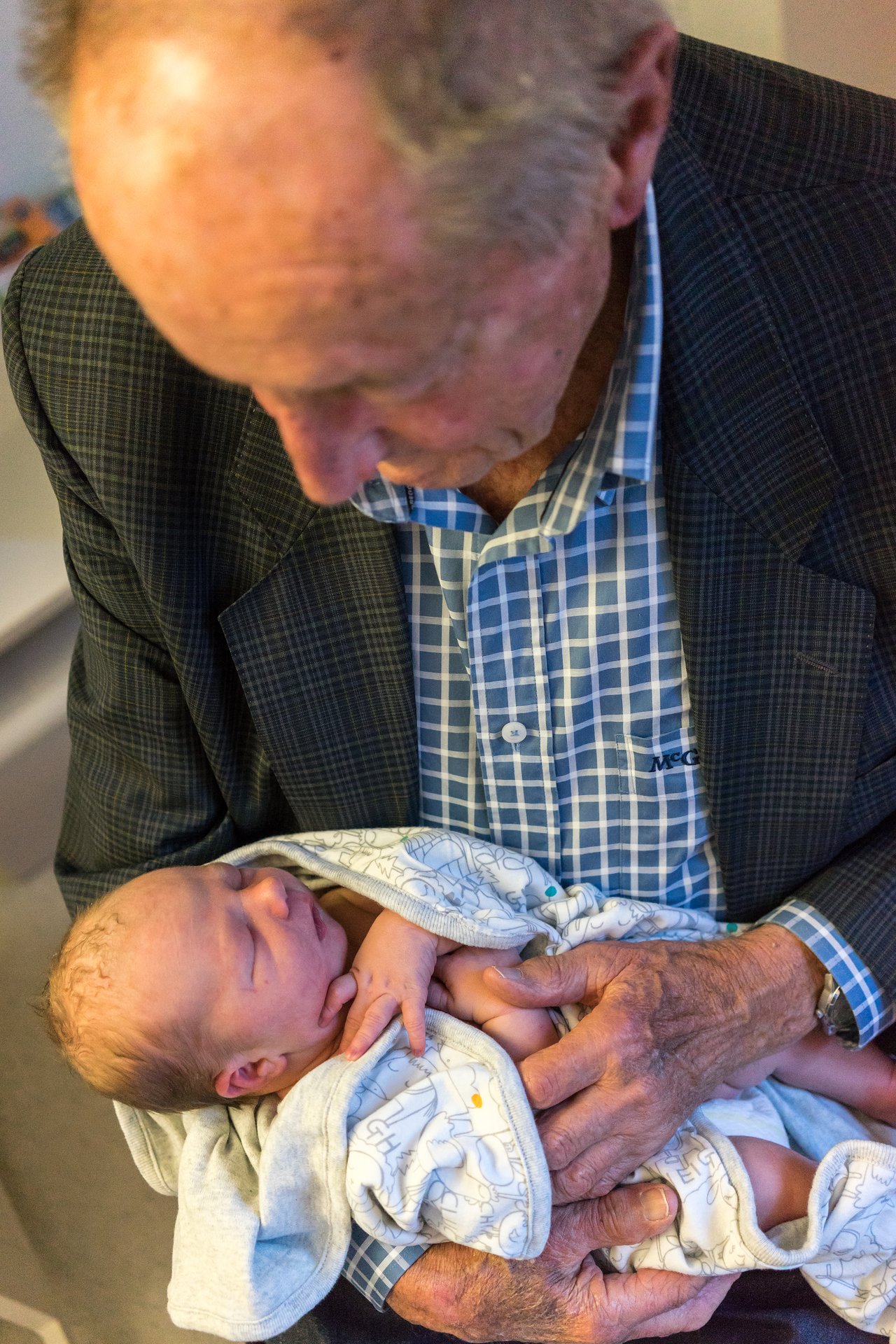 An elderly man in a suit holds a newborn baby wrapped in a blanket, looking down at the child.