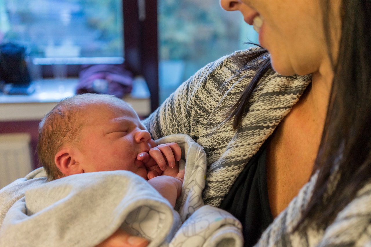 A woman smiles while holding a newborn baby wrapped in a blanket, who is sleeping with a hand near their mouth.