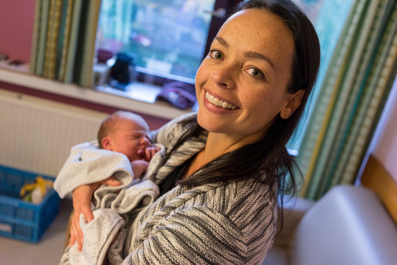 A smiling woman holds a newborn baby wrapped in a blanket, looking at the camera.