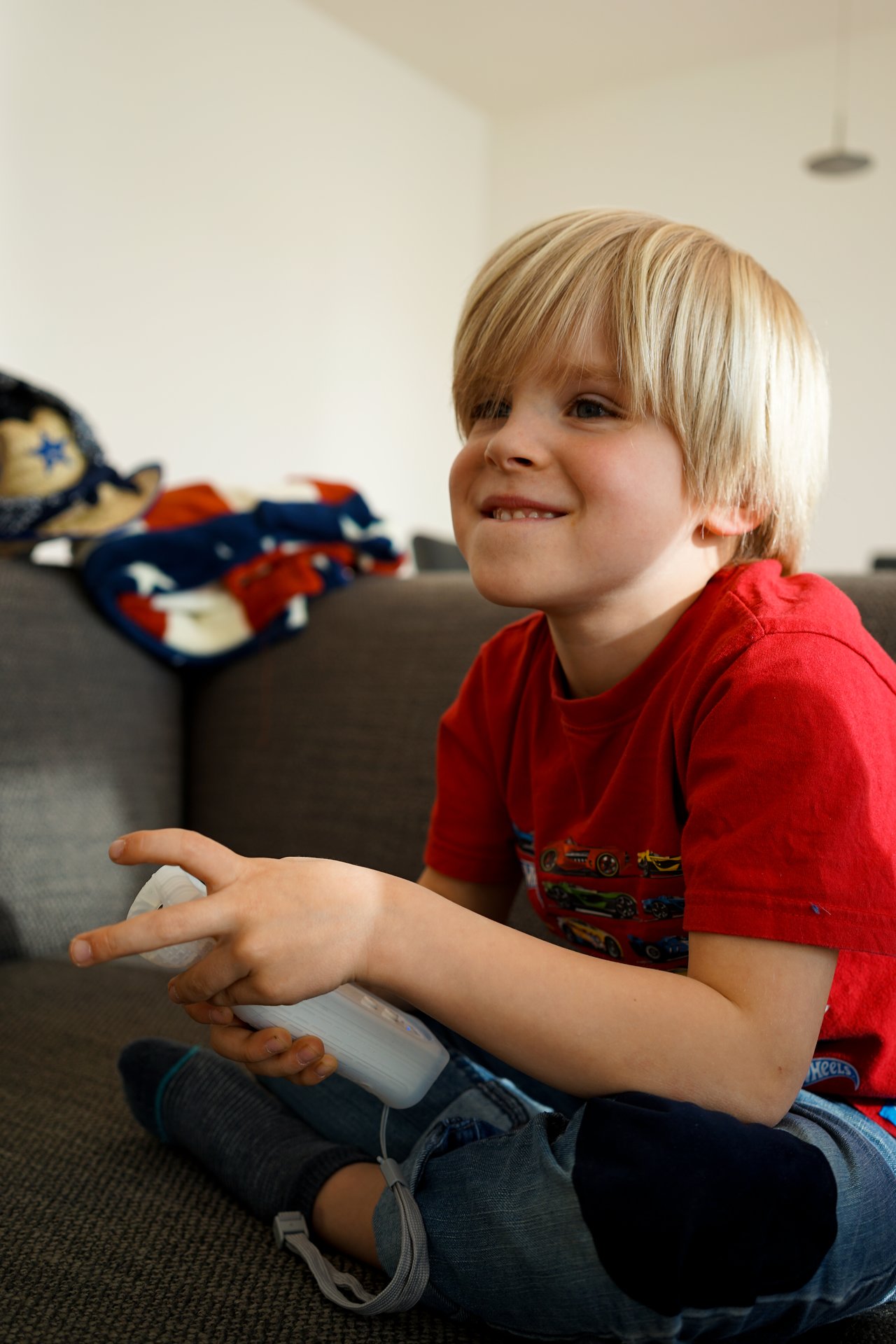 A young child in a red shirt sits on a couch, smiling while playing a Wii game with a controller.