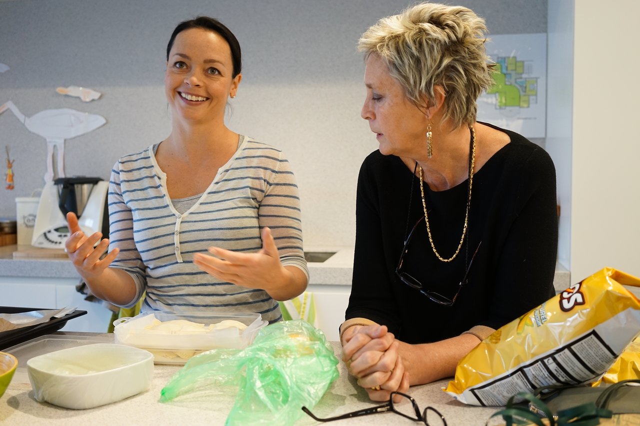 A woman in a striped shirt talks and gestures while another woman in black listens at a kitchen counter.