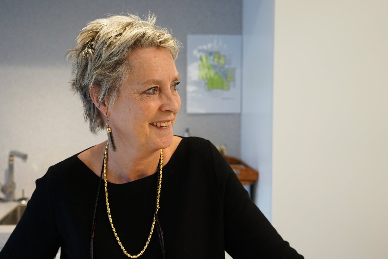 An older woman with short gray hair and a black top smiles while looking to the side in a kitchen.