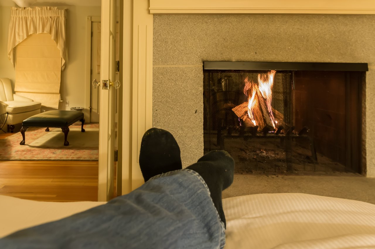 Person relaxing on a bed with feet up, facing a lit fireplace in a cozy room.