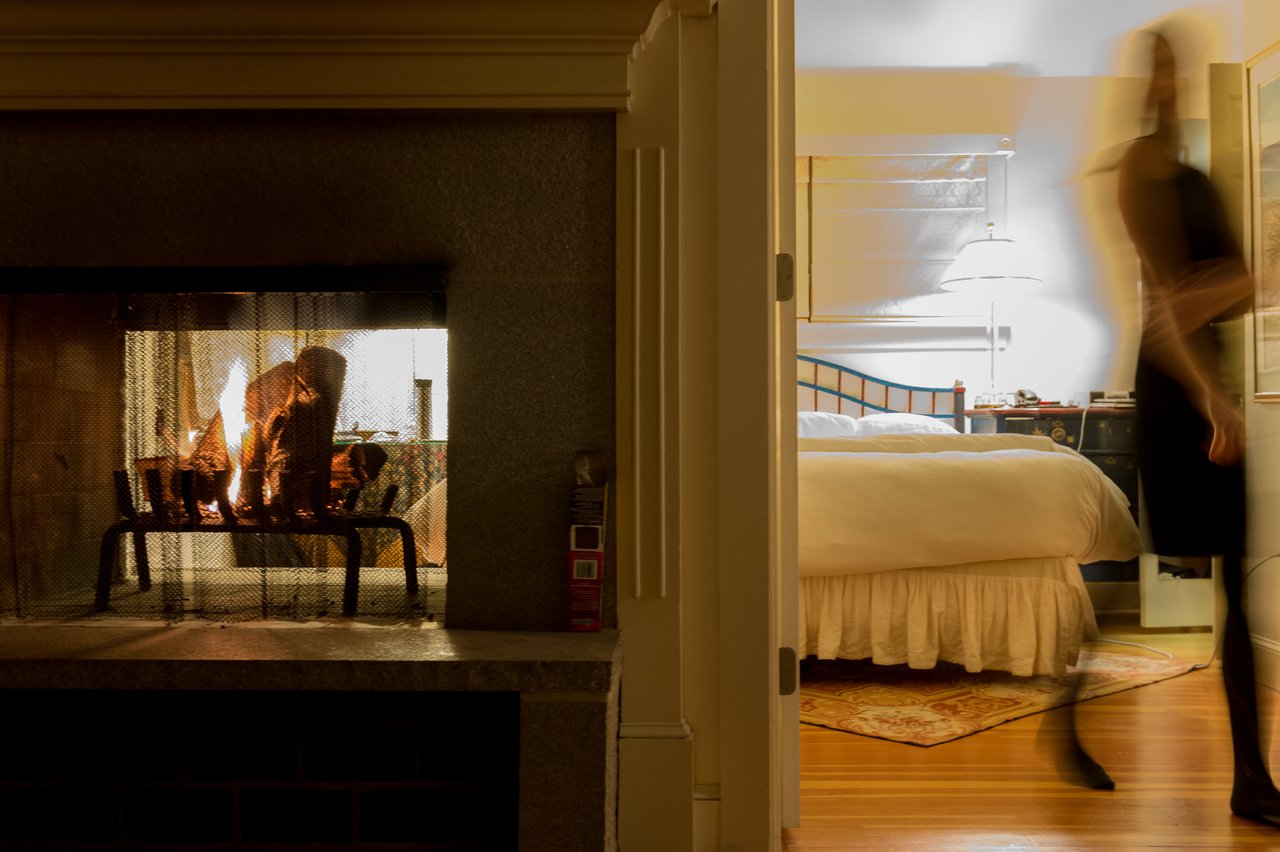 A fireplace with burning logs on the left, and a blurred person walking past a bedroom on the right.