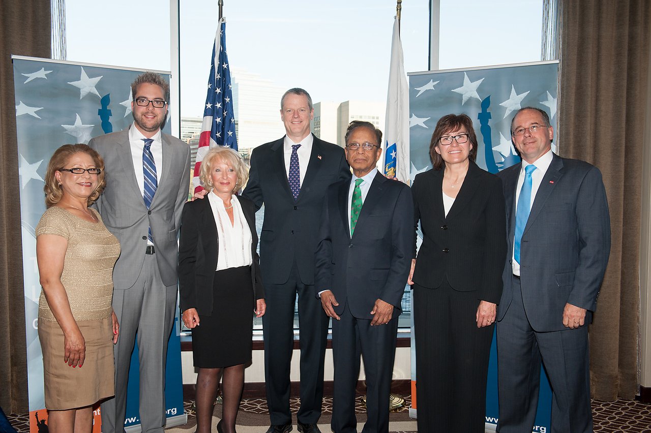 A group of seven professionally dressed people pose for a photo in front of flags and banners at an event.