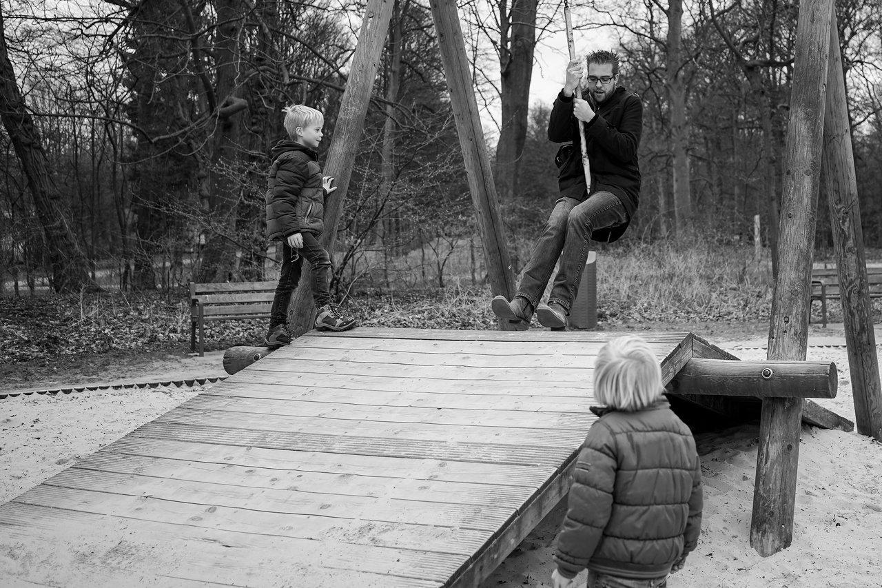 A man rides a zipline at a playground while two children watch from the wooden platform.
