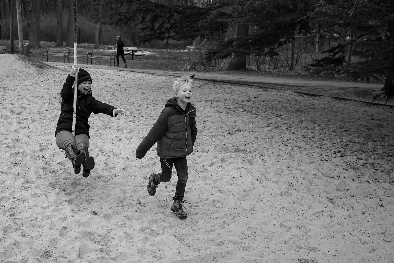 A child rides a zipline while another child runs alongside, both smiling and enjoying the playground.