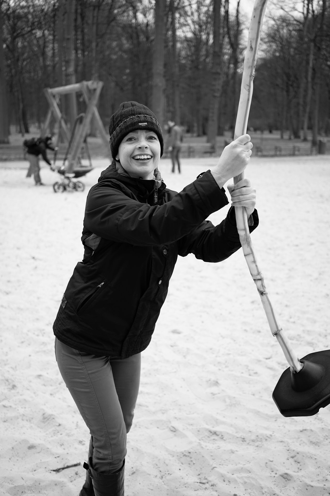 A person in a winter jacket and hat holds onto a zipline handle, smiling in an outdoor park.