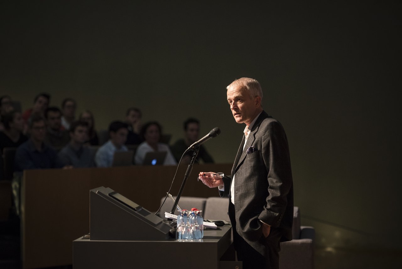 A university official stands at a podium, speaking into a microphone while addressing an audience in a lecture hall.