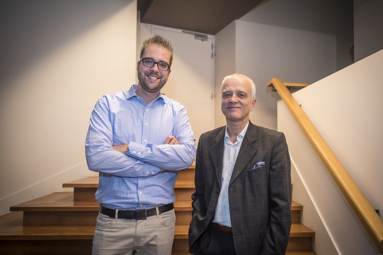 Two men standing side by side, smiling at the camera, in front of a staircase indoors.