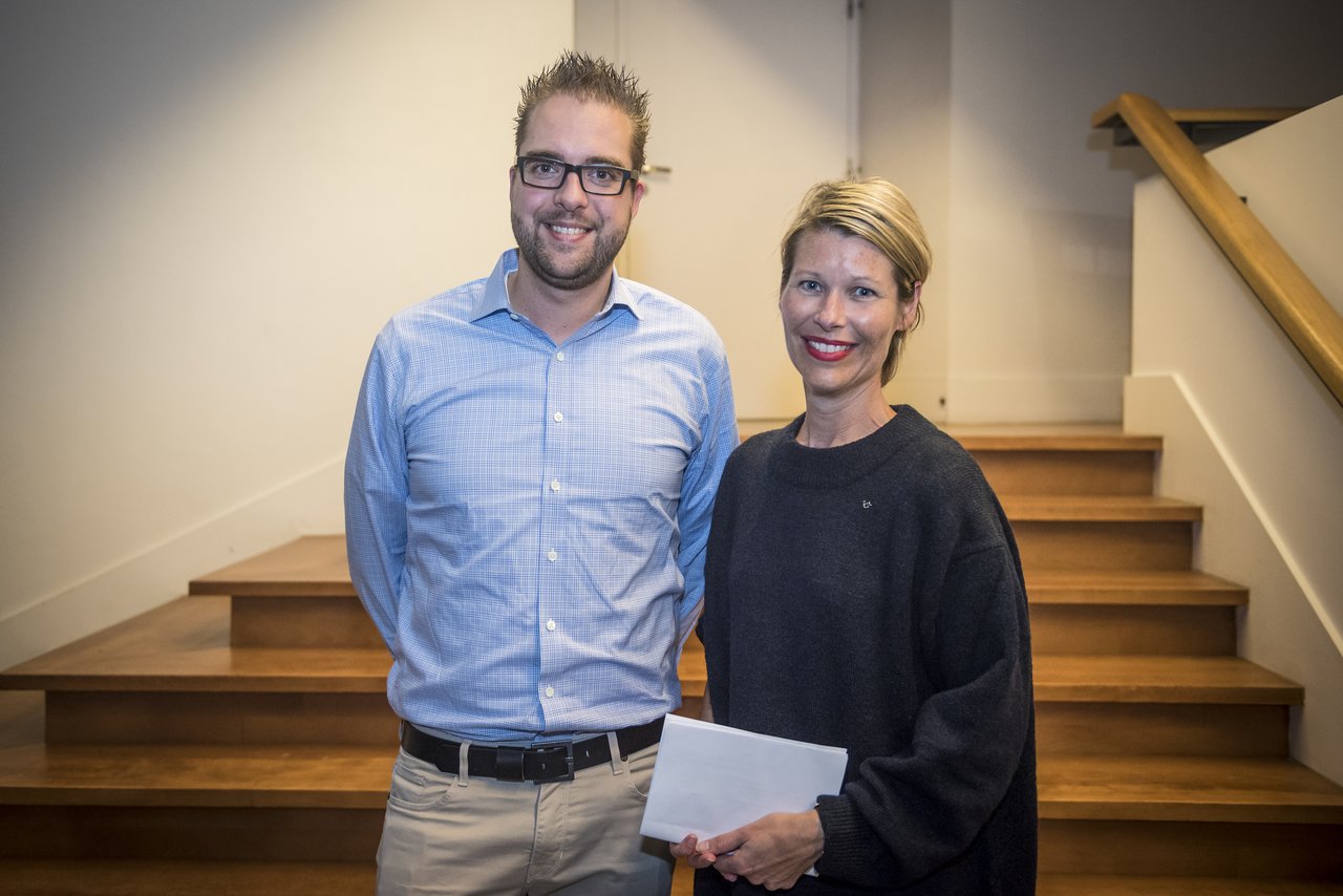 Two people stand in front of wooden stairs, smiling.
