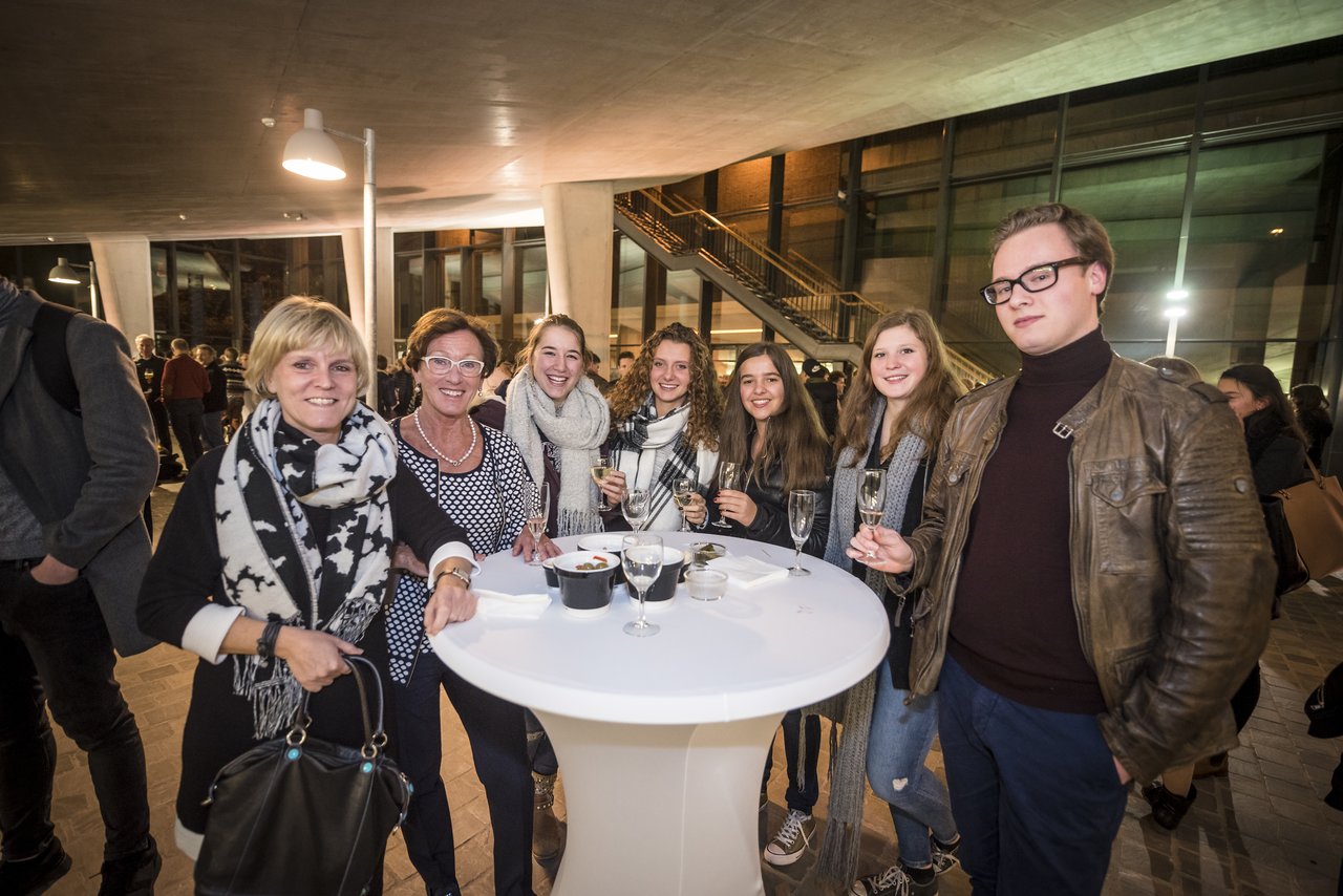 A group of people stands around a table, holding drinks and smiling at a university presentation event in Antwerp.