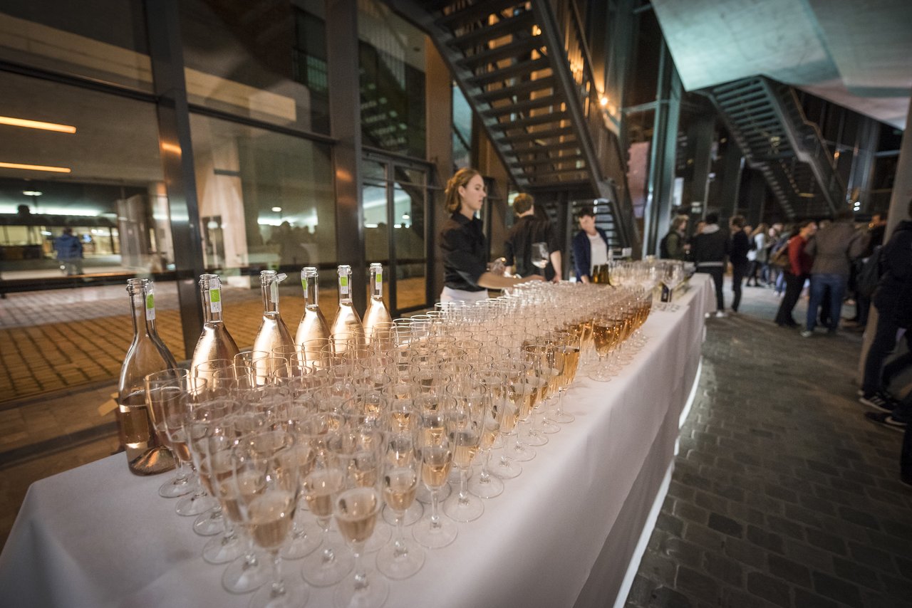 A server arranges champagne glasses on a table at a university event, with guests gathering in the background.