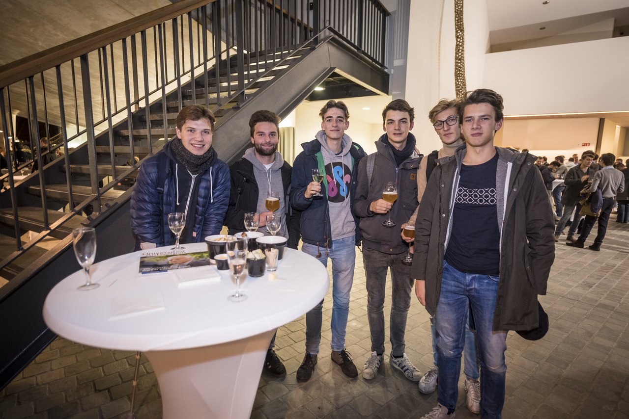 A group of young men stand together at a university event, holding drinks and smiling near a table with refreshments.