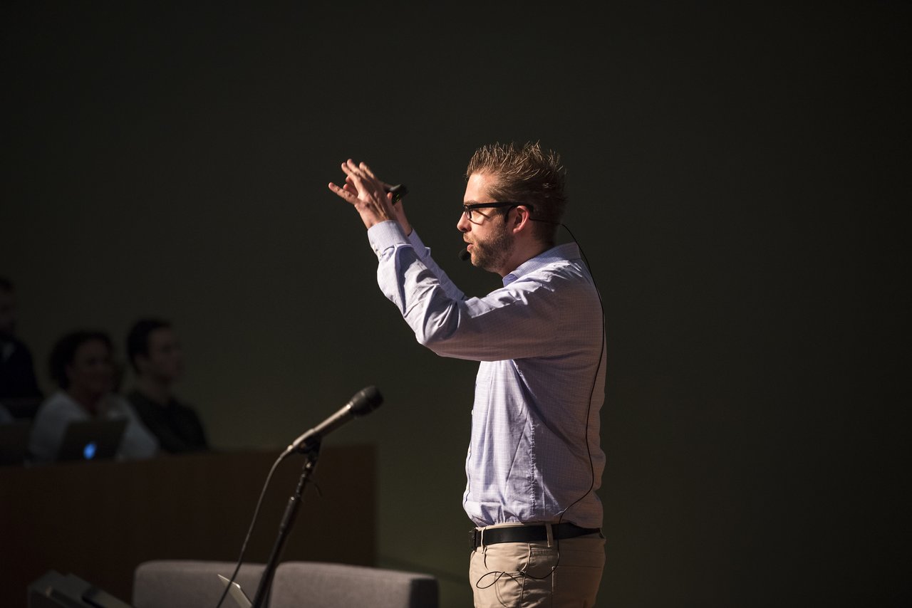 A speaker in a light blue shirt gestures while presenting at Antwerp University, with a microphone nearby.