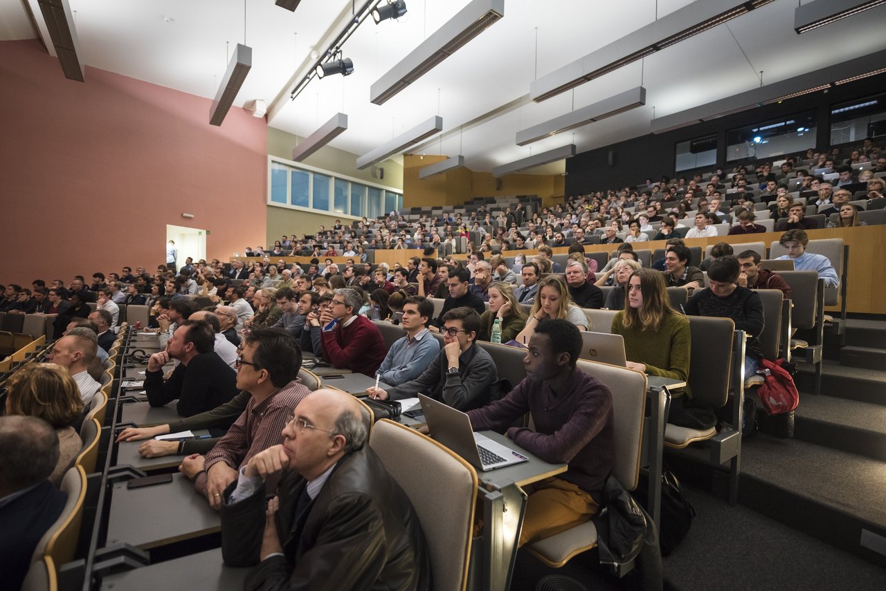 A large audience in a university lecture hall attentively listens to a presentation, with some taking notes on laptops.